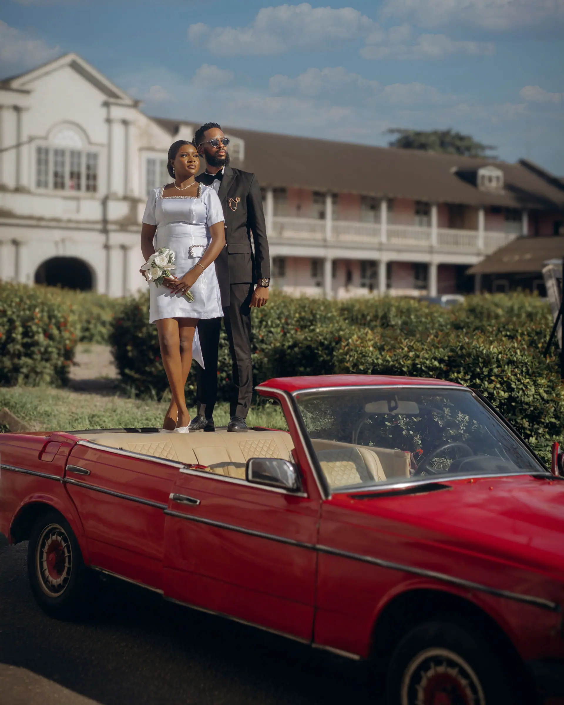 Amara and Ebuka standing on a red convertible outside a colonial building in Lagos
